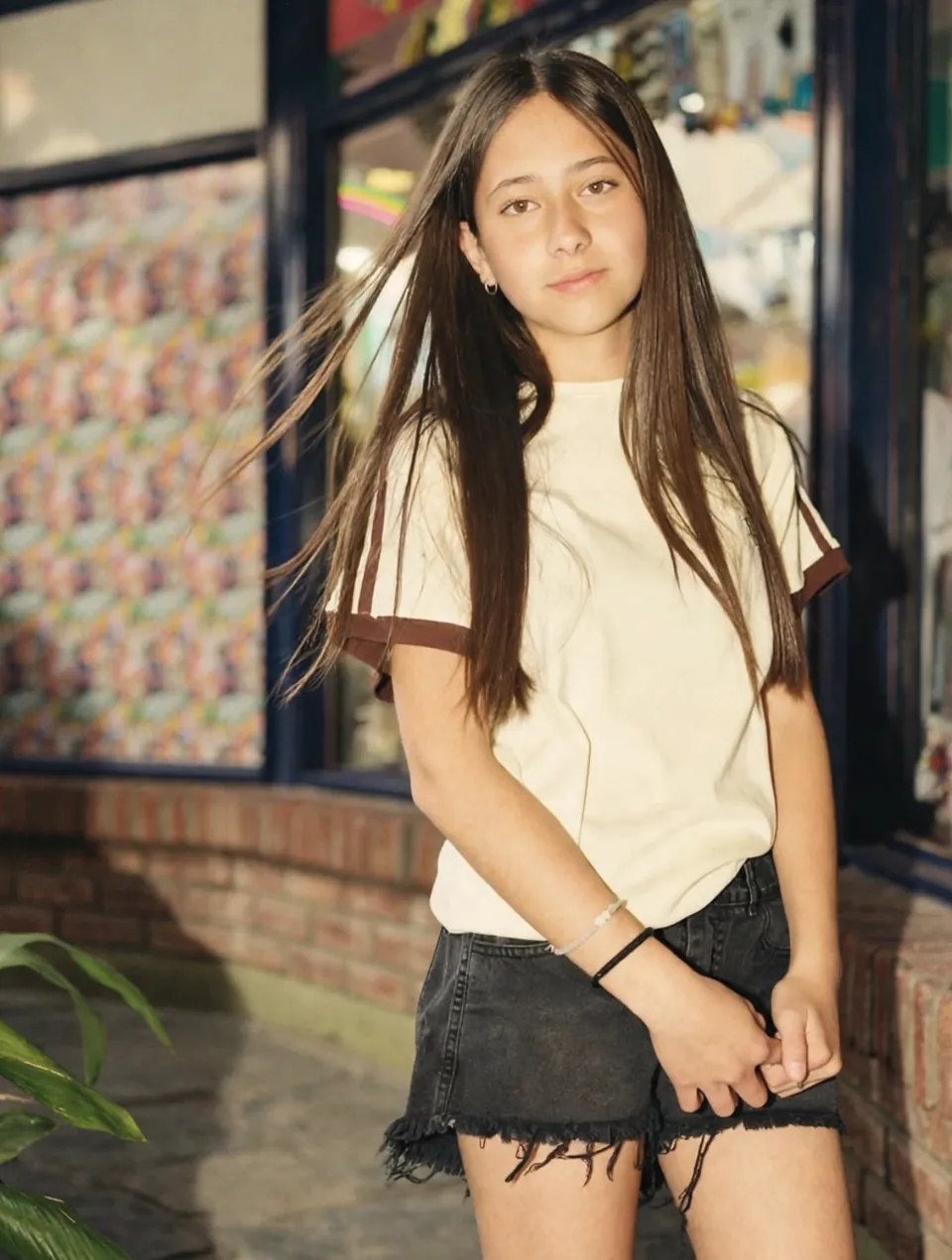 Chica de pelo largo con remera crema de mangas marrones y short negro, frente a una vidriera
